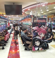 A bustling arcade filled with people playing various gaming machines. Brightly colored displays and animated characters are prominent on posters and screens. The atmosphere is lively with patrons seated in rows, focusing on the games. Overhead signage and electronic scoreboards are visible, contributing to the busy and vibrant environment.
