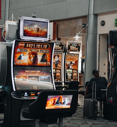 A collection of slot machines is situated inside a building with bright, colorful screens displaying various games and gaming themes. A person is seated nearby, seemingly relaxed with luggage beside them, and the atmosphere is lively with electronic displays.